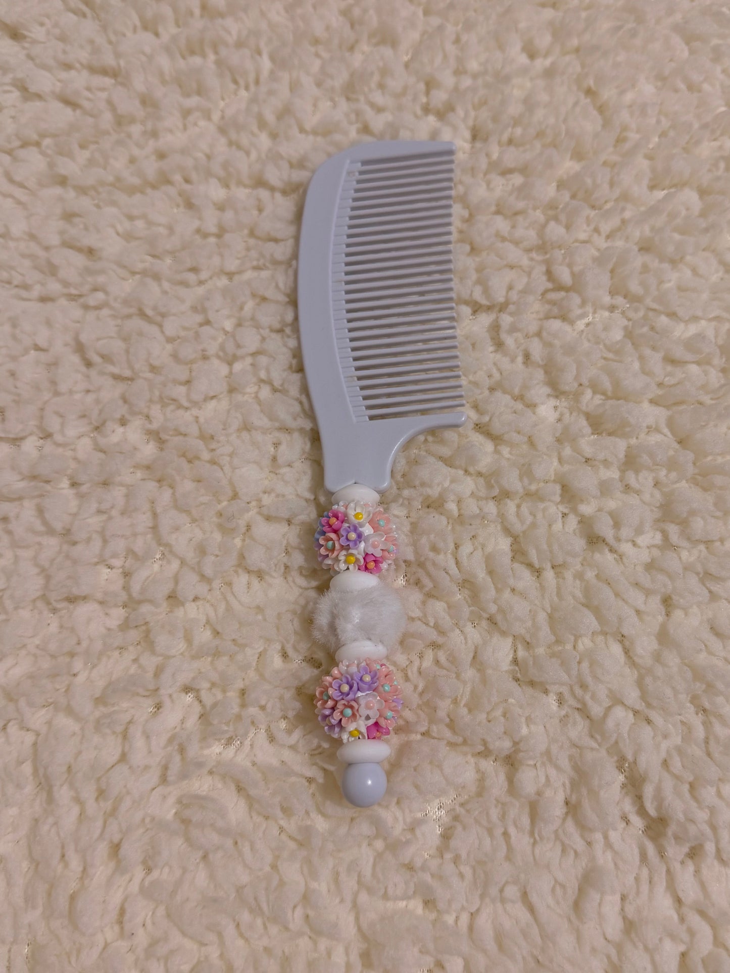 A light gray plastic hair comb with decorative white and multicolor beaded handle details, placed on a textured cream-colored fleece surface.