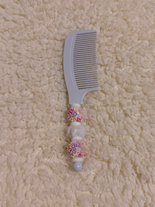 A light gray plastic hair comb with decorative white and multicolor beaded handle details, placed on a textured cream-colored fleece surface.
